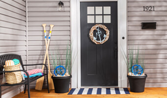 Black planters filled with seashells, tall blades of grass and a blue, decorative metal orb sit on either side of the front door. A round twine wreath with a large blue anchor in the center hangs on the front door. 