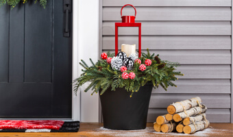 A black planter filled with greenery, painted pine cones and a red lantern holding a white candle. The planter is on a front porch to the right of the front door. 