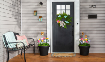 Black planters filled with greenery, painted wooden flowers and small, wood stained bird houses sit on either side of the front door. A green wreath decorated with painted flowers and birds hangs on the front door.