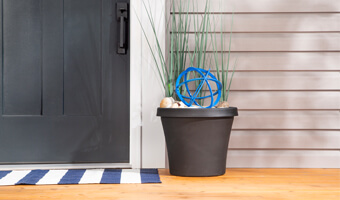 A black planter filled with seashells, tall blades of grass and a blue, decorative metal orb. The planter is on a front porch to the right of the front door. 
