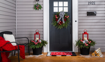 Black planters filled with greenery, painted pine cones and a red lantern holding a white candle sit on either side of the front door. An evergreen wreath decorated with painted pine cones and a black and white bow is hanging on the front door. 