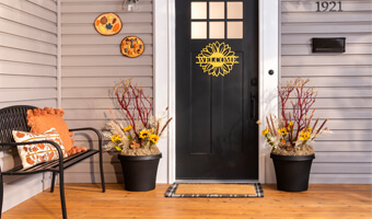 A front porch with black planters filled with decorative sunflowers and manzanita branches on either side of the front door. A yellow welcome sign in the shape of a sunflower is hanging on the front door. 