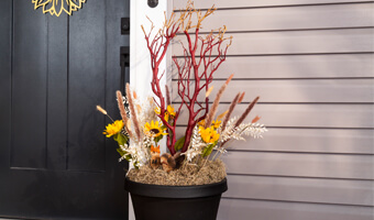 A black planter filled with decorative sunflowers and manzanita branches. The planter is on a front porch to the right of the front door. 
